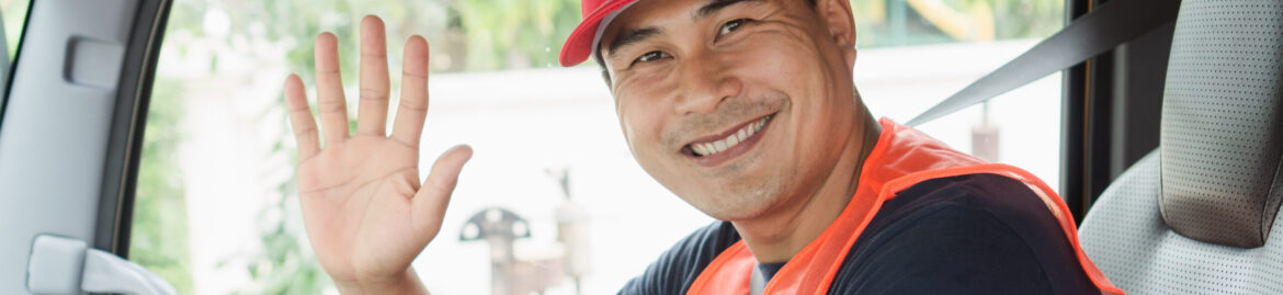 A smiling truck driver in a red cap and high-visibility vest, seated in the driver's seat, waving.