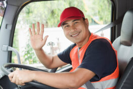 A smiling truck driver in a red cap and high-visibility vest, seated in the driver's seat, waving.